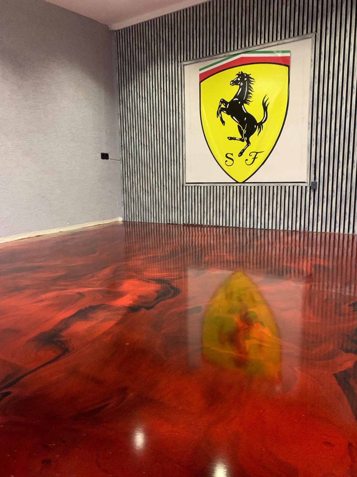 Glänzender roter Epoxidharzboden mit schwarzen Strukturen vor einer Lamellenwand mit Ferrari-Emblem